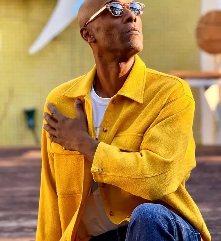 Marcus Chocollate in a yellow jacket and sunglasses, hand on his chest, looking up.